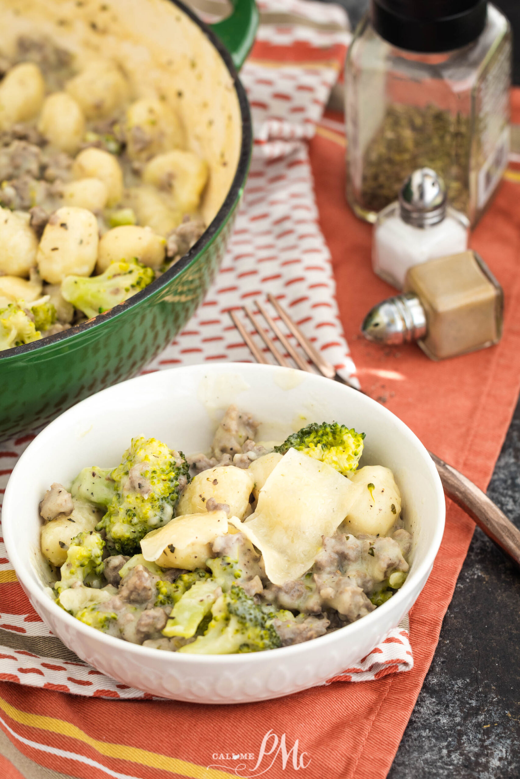 A white bowl filled with gnocchi, ground meat, broccoli, and sauce sits on a patterned napkin next to a fork, with a green pot and seasonings in the background.