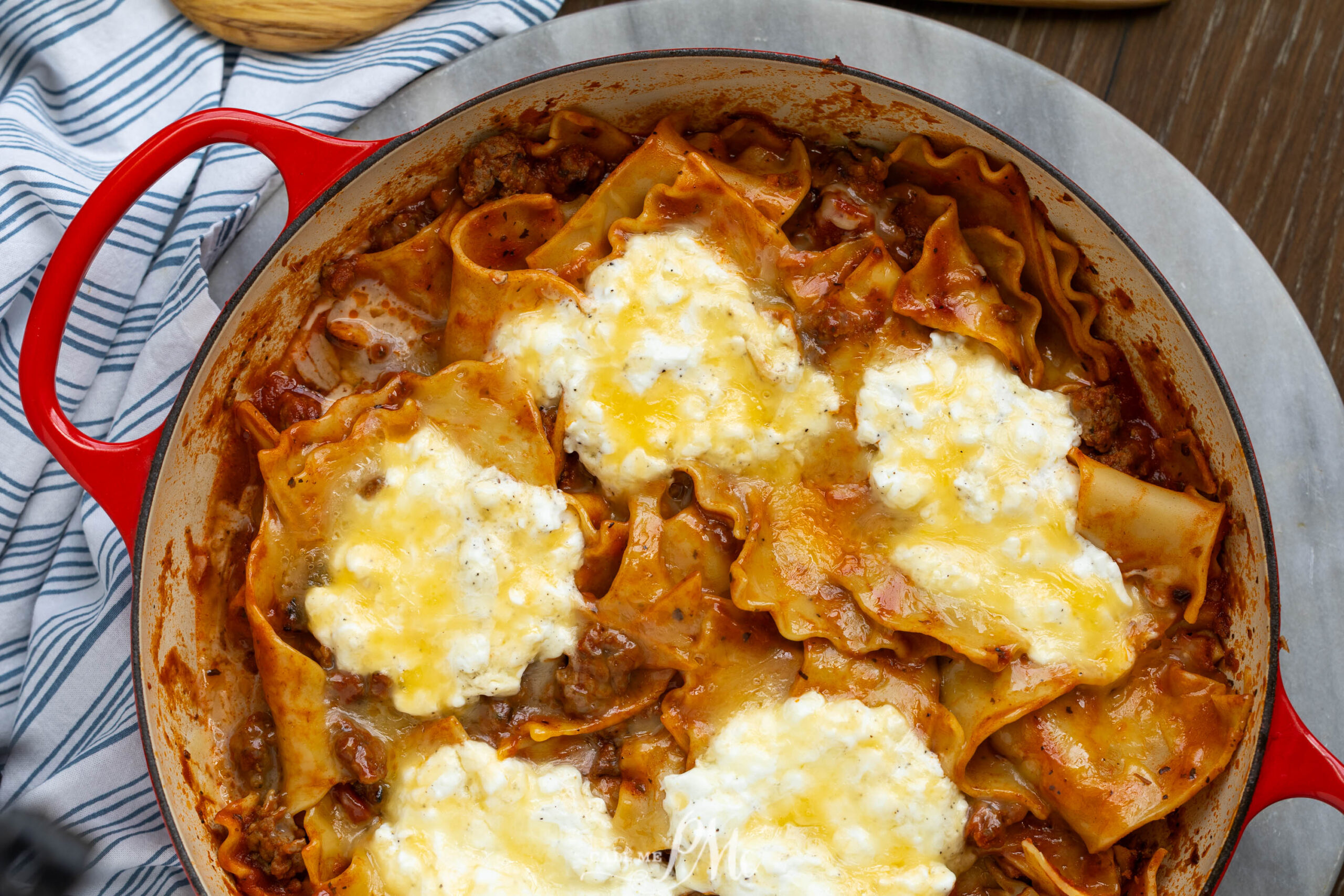 A red skillet filled with baked lasagna, topped with melted cheese and dollops of ricotta, sits on a blue-striped cloth and silver tray.