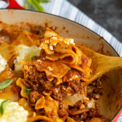 A wooden spoon lifts a serving of cheesy, meaty lasagna from a red casserole dish, with layers of pasta, sauce, and ground beef visible.