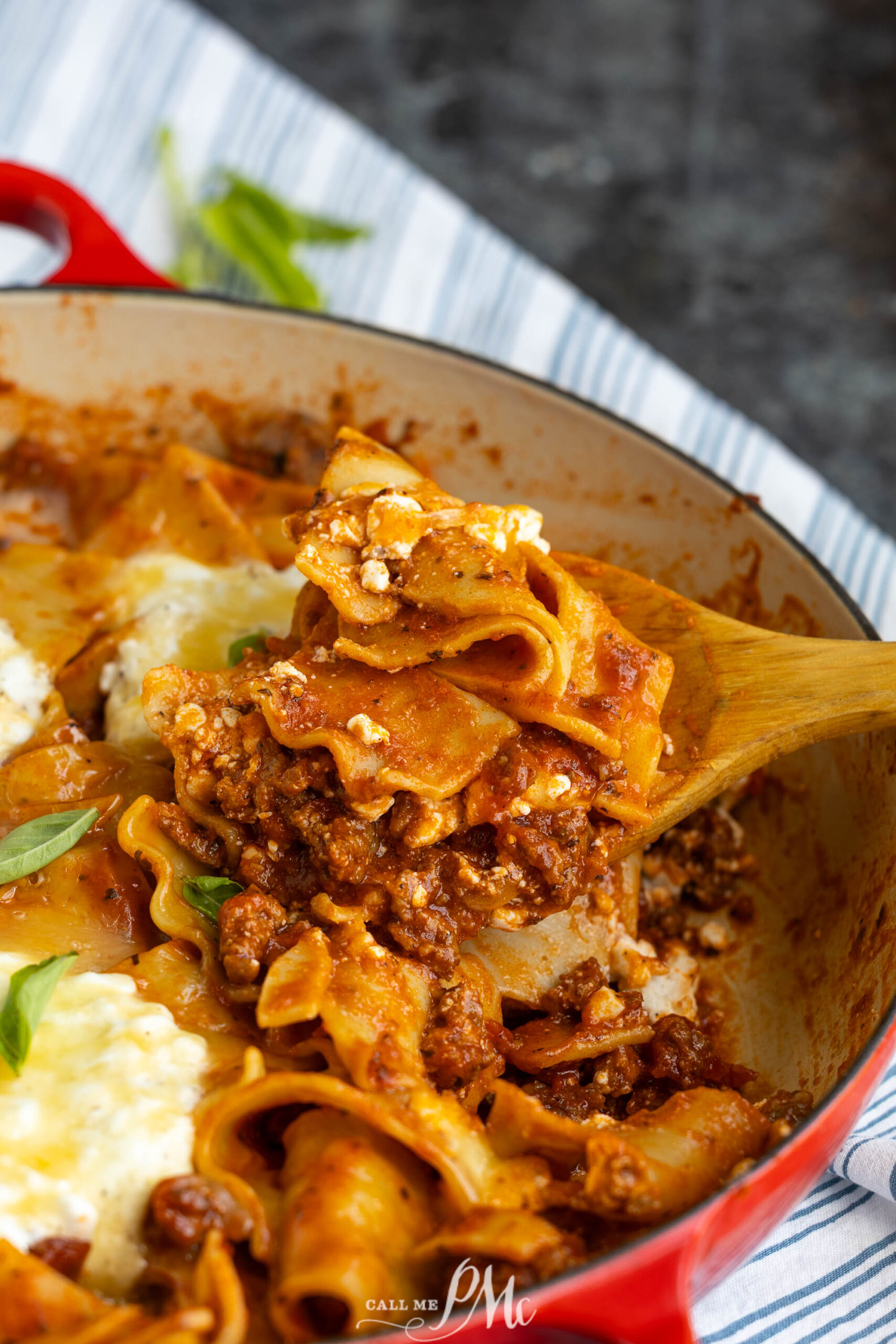 A wooden spoon lifts a serving of cheesy, meaty lasagna from a red casserole dish, with layers of pasta, sauce, and ground beef visible.