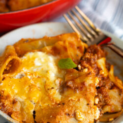 A close-up of a plate of lasagna with melted cheese and meat sauce, sitting on a striped napkin next to a fork. A red baking dish with more lasagna is in the background.