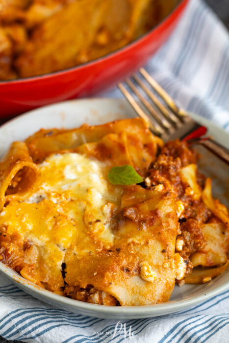 A close-up of a plate of lasagna with melted cheese and meat sauce, sitting on a striped napkin next to a fork. A red baking dish with more lasagna is in the background.
