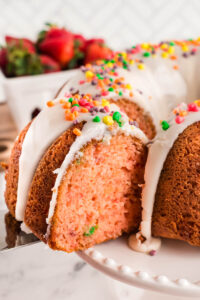 A close-up of a Nerds Strawberry Bundt Cake with white icing and colorful sprinkles, with a slice being lifted out. Fresh strawberries are blurred in the background.