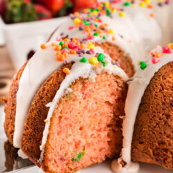 A close-up of a Nerds Strawberry Bundt Cake with white icing and colorful sprinkles, with a slice being lifted out. Fresh strawberries are blurred in the background.