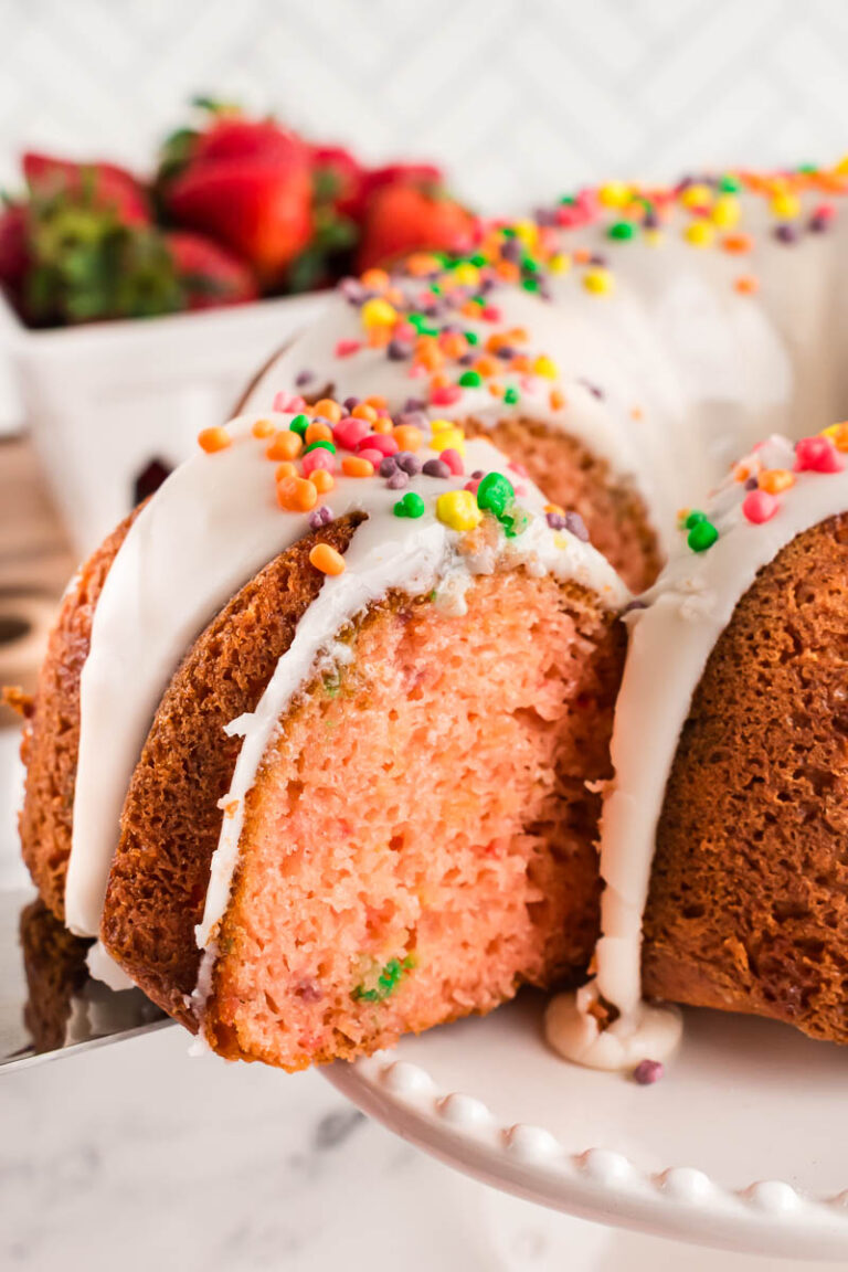 A close-up of a Nerds Strawberry Bundt Cake with white icing and colorful sprinkles, with a slice being lifted out. Fresh strawberries are blurred in the background.