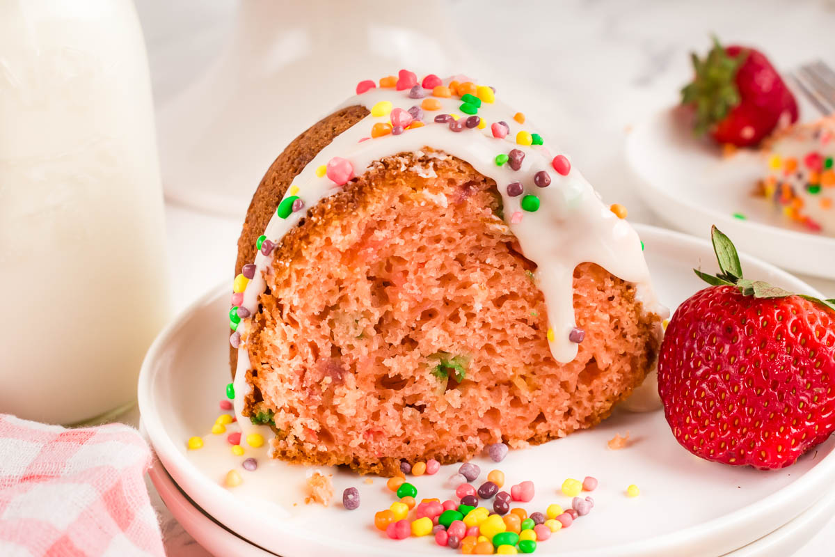 A slice of dessert with white icing and colorful round sprinkles sits on a plate next to a fresh strawberry.