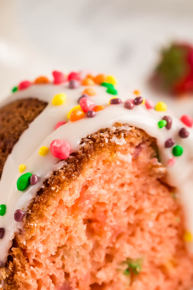 Close-up of a slice of Nerds Strawberry Bundt Cake with white icing and colorful round sprinkles, showing a moist texture. A blurred strawberry is visible in the background.