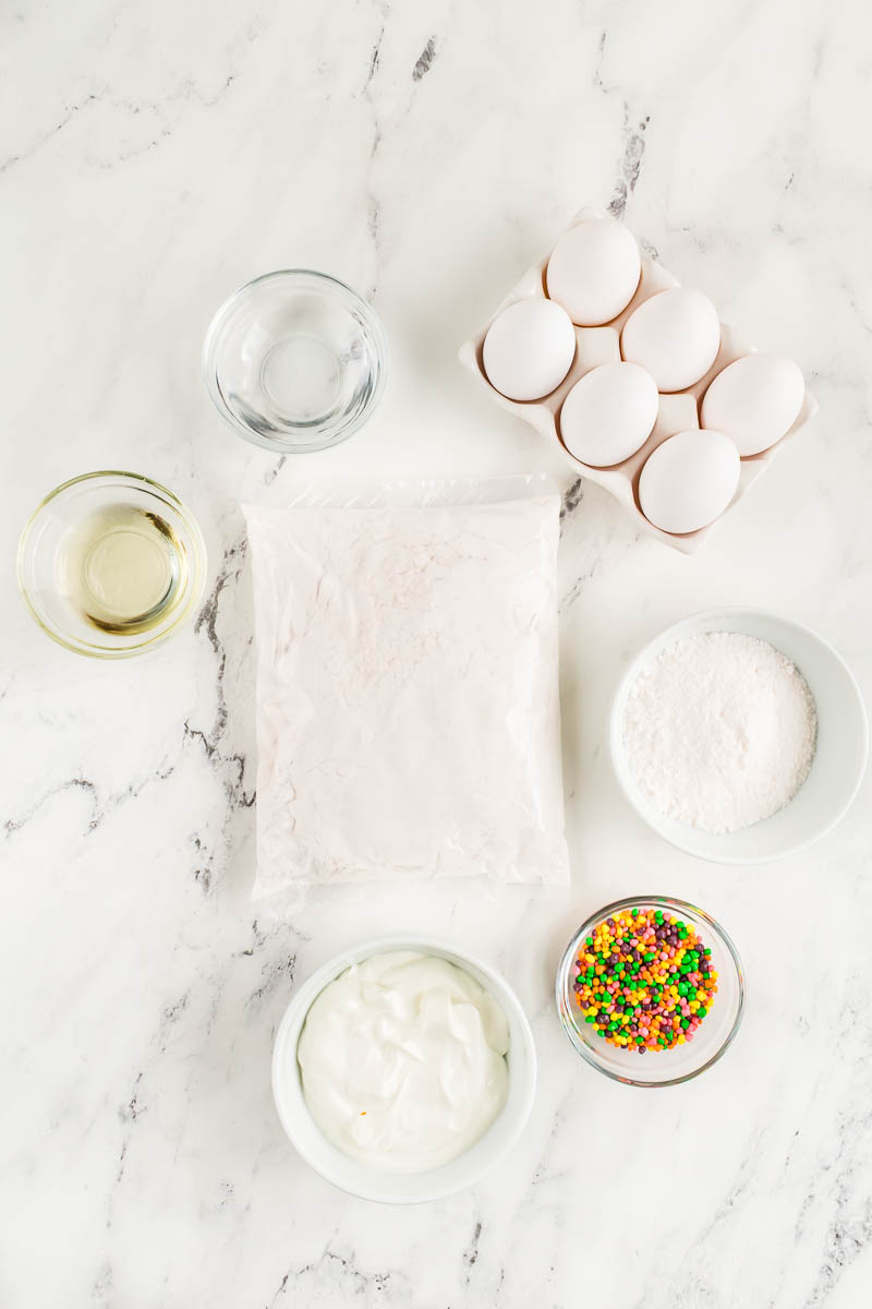 A flat lay of baking ingredients: a pack of cake mix, six eggs in a carton, a bowl of yogurt, a bowl of flour, a bowl of sprinkles, a bowl of oil, and a bowl of water arranged on a white marble surface.