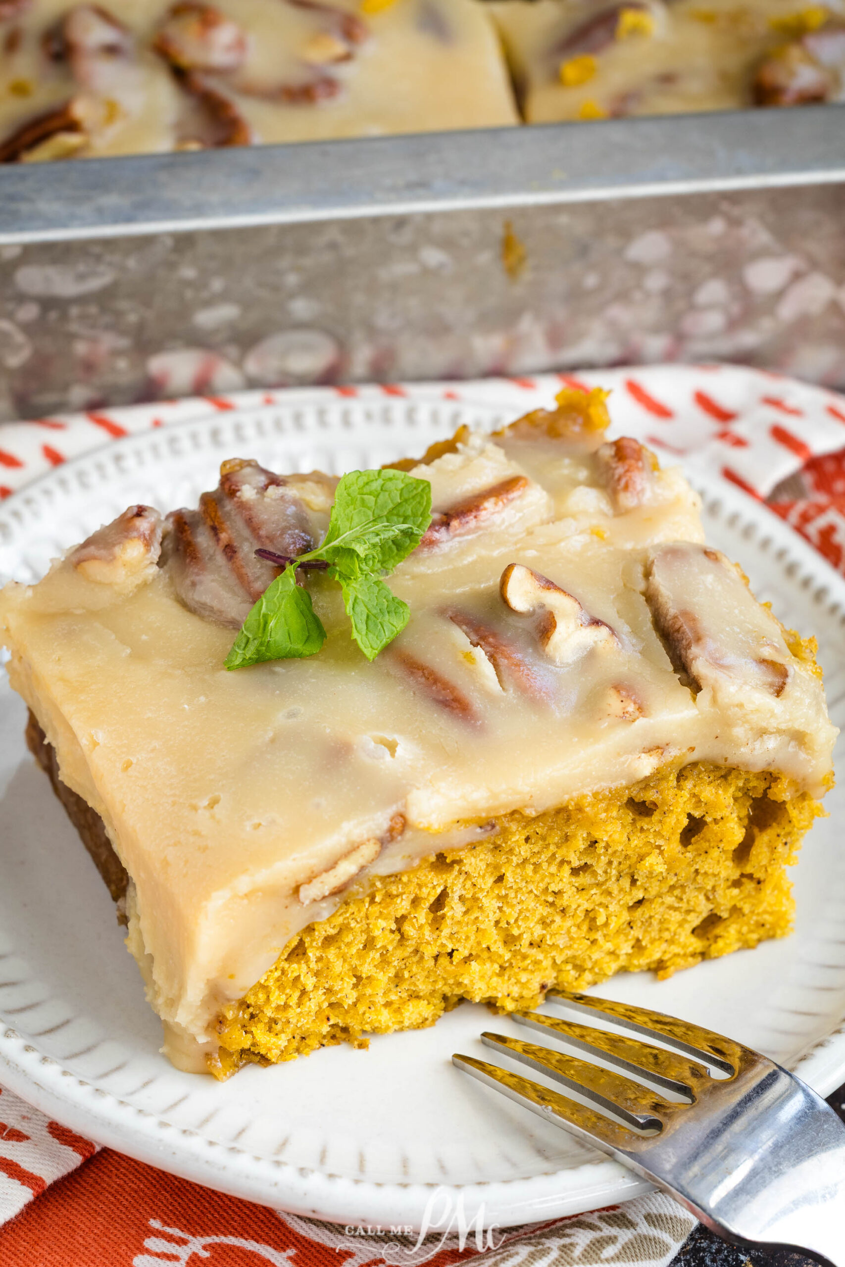 A slice of pumpkin cake topped with creamy icing and pecans sits on a white plate beside a fork, with a sprig of mint for garnish. The cake pan and more cake are visible in the background.