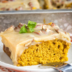 A slice of moist pumpkin cake with creamy frosting and pecans sits on a white plate with a fork, garnished with a sprig of mint. The rest of the cake is visible in a pan in the background.