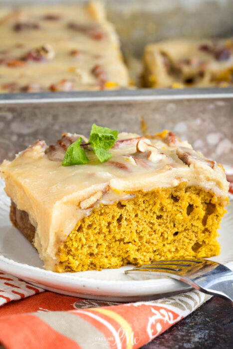 A slice of moist pumpkin cake with creamy frosting and pecans sits on a white plate with a fork, garnished with a sprig of mint. The rest of the cake is visible in a pan in the background.
