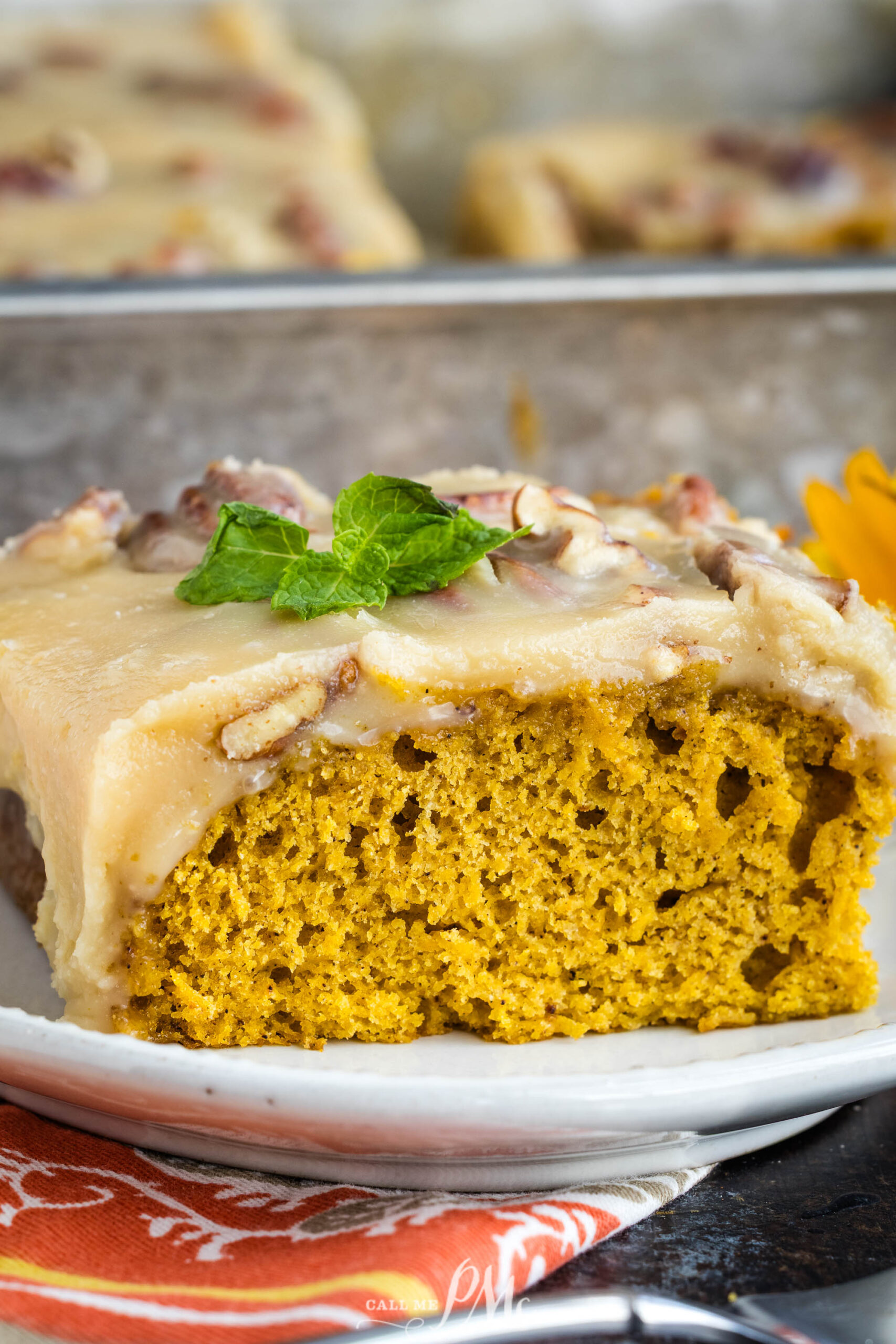 A slice of moist dessert with light brown frosting garnished with a mint leaf, sits on a white plate with a colorful napkin underneath.
