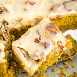 A close-up of pumpkin cake bars topped with creamy frosting and pecans, one bar lifted from the tray with a spatula, showing moist texture and scattered crumbs.