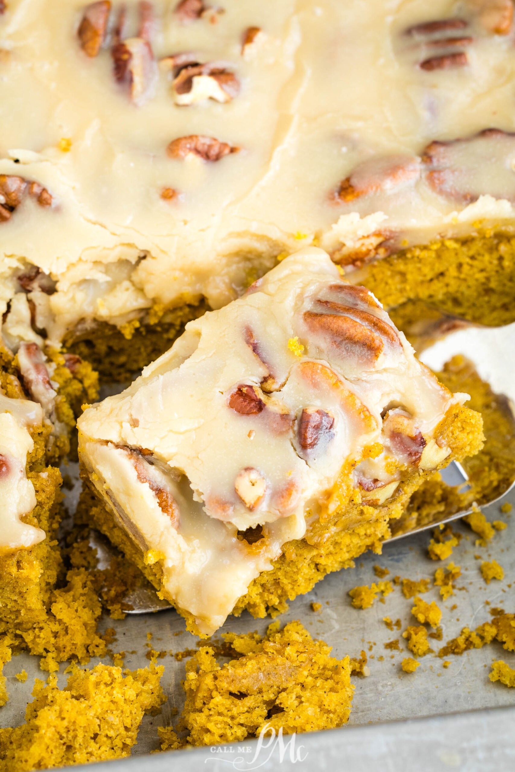 A close-up of pumpkin cake bars topped with creamy frosting and pecans, one bar lifted from the tray with a spatula, showing moist texture and scattered crumbs.