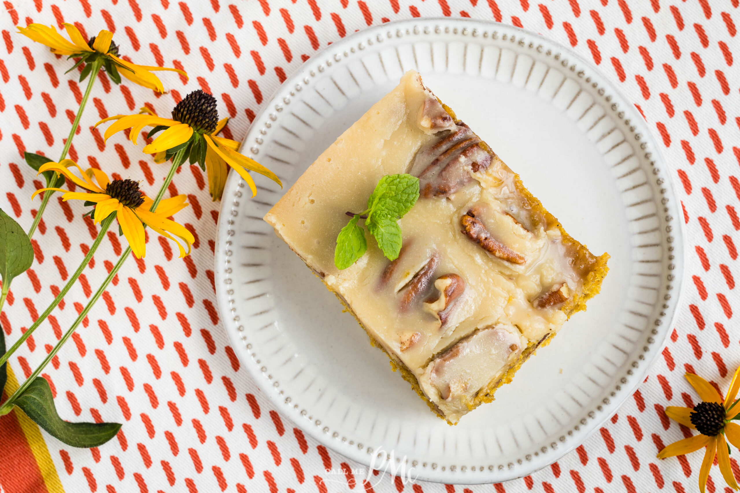 A slice of pecan-topped cake with creamy icing, garnished with mint, sits on a white plate. Yellow flowers and a red-patterned cloth surround the plate.