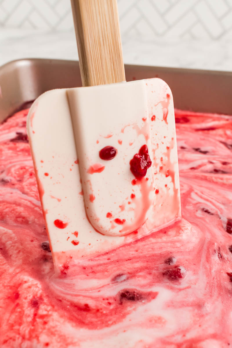 A white spatula with a wooden handle stirs a pink and red swirled creamy mixture with berry pieces in a metal pan.