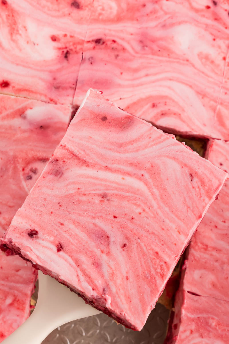 A close-up of a square raspberry swirl cheesecake bar, its pink and white marbled patterns reminiscent of Raspberry Snow Salad, being lifted from a tray of similar bars.