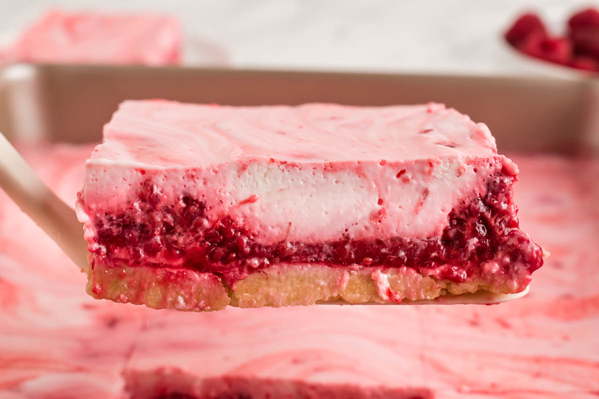 A close-up of a dessert bar with a cookie crust, a red berry filling and a pink and white creamy layer on top, being lifted from a pan.