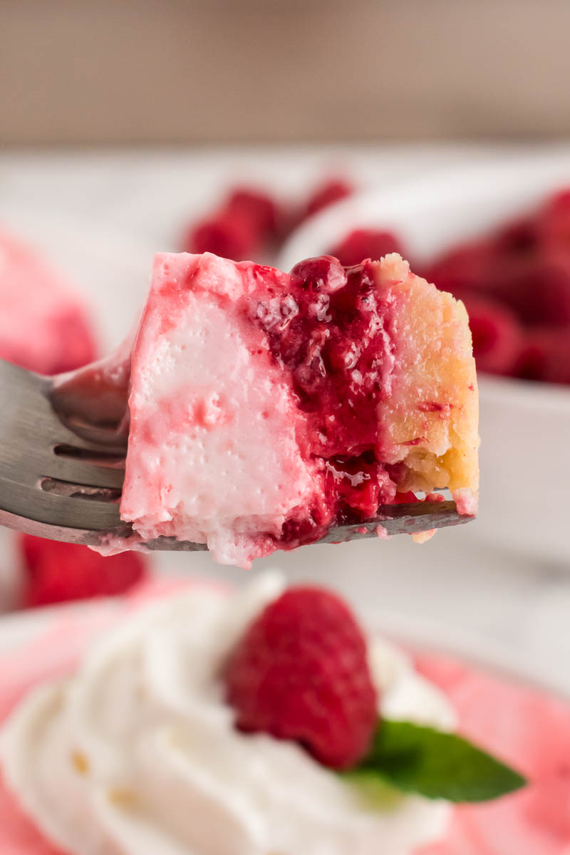 A fork holds a creamy pink and white Raspberry Snow Salad dessert with a crumbly crust and raspberry topping, with whipped cream, a raspberry, and a mint leaf blurred in the background.