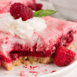 A slice of Raspberry Snow Salad dessert with whipped cream, a raspberry, and a mint leaf on top sits on a plate. A bite has been taken out, and fresh raspberries are scattered nearby.