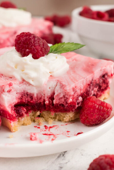 A slice of Raspberry Snow Salad dessert with whipped cream, a raspberry, and a mint leaf on top sits on a plate. A bite has been taken out, and fresh raspberries are scattered nearby.