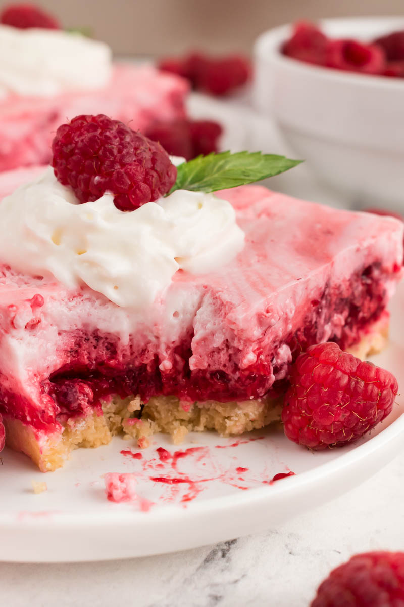 A slice of Raspberry Snow Salad dessert with whipped cream, a raspberry, and a mint leaf on top sits on a plate. A bite has been taken out, and fresh raspberries are scattered nearby.