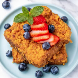 Two slices of cereal-crusted French toast topped with sliced strawberries and a mint sprig, surrounded by fresh blueberries on a light blue plate.