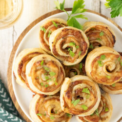 A white plate piled with golden-brown savory ham and cheese pinwheels topped with fresh herbs.