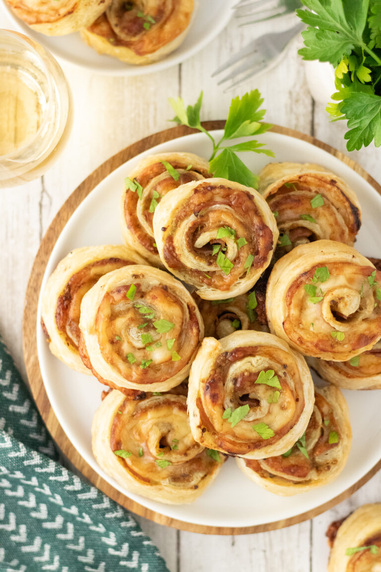 A white plate piled with golden-brown savory ham and cheese pinwheels topped with fresh herbs.