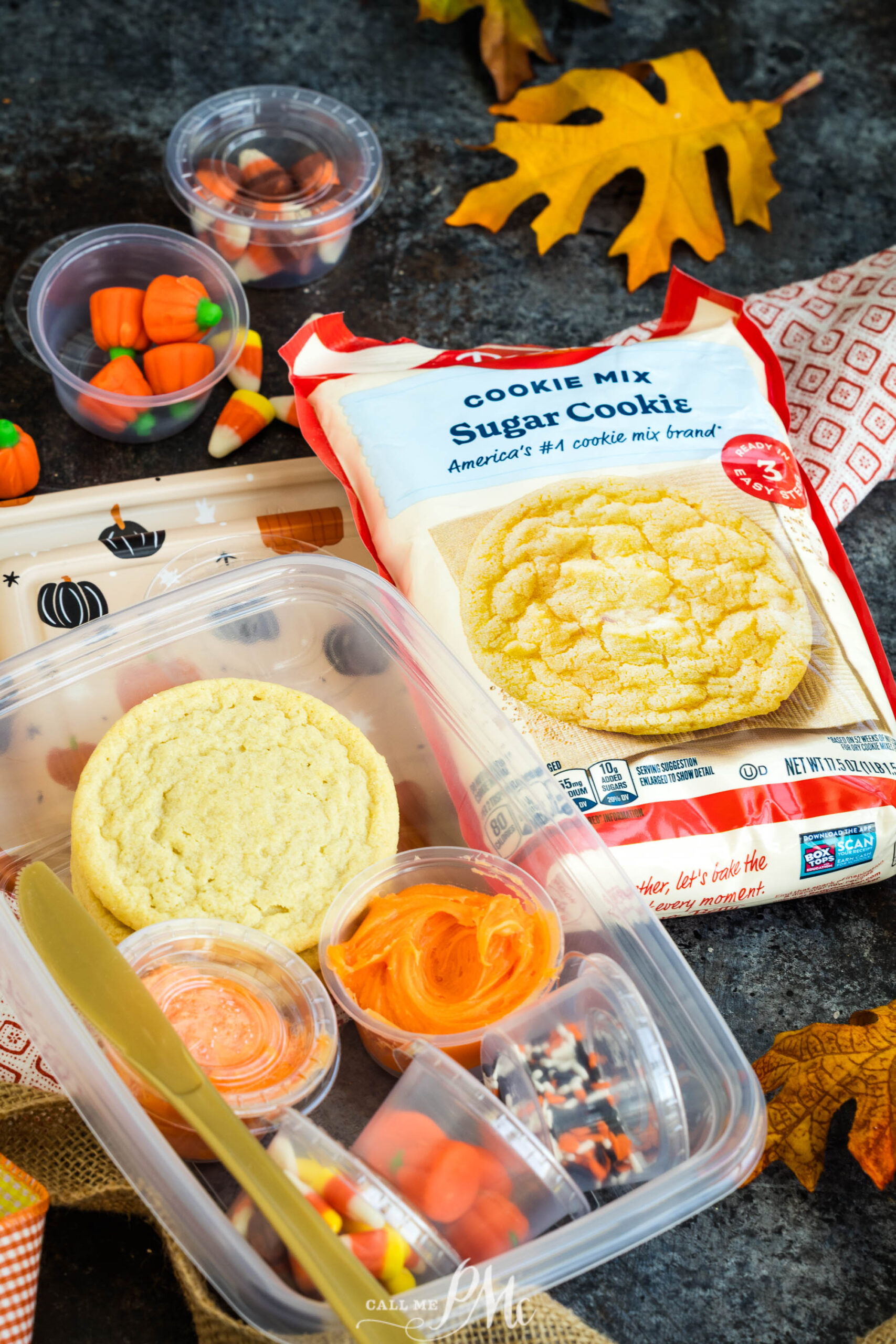A DIY Halloween cookie kit consisting of a sugar cookie mix pouch next to a plastic container holding a baked cookie, orange frosting, and containers of themed candy.
