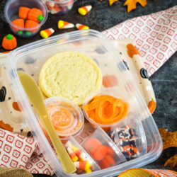 A plastic lunch container holding a cookie, orange icing, candy corn, sprinkles, and gummy pumpkins, with a plastic knife and surrounding fall-themed decorations.