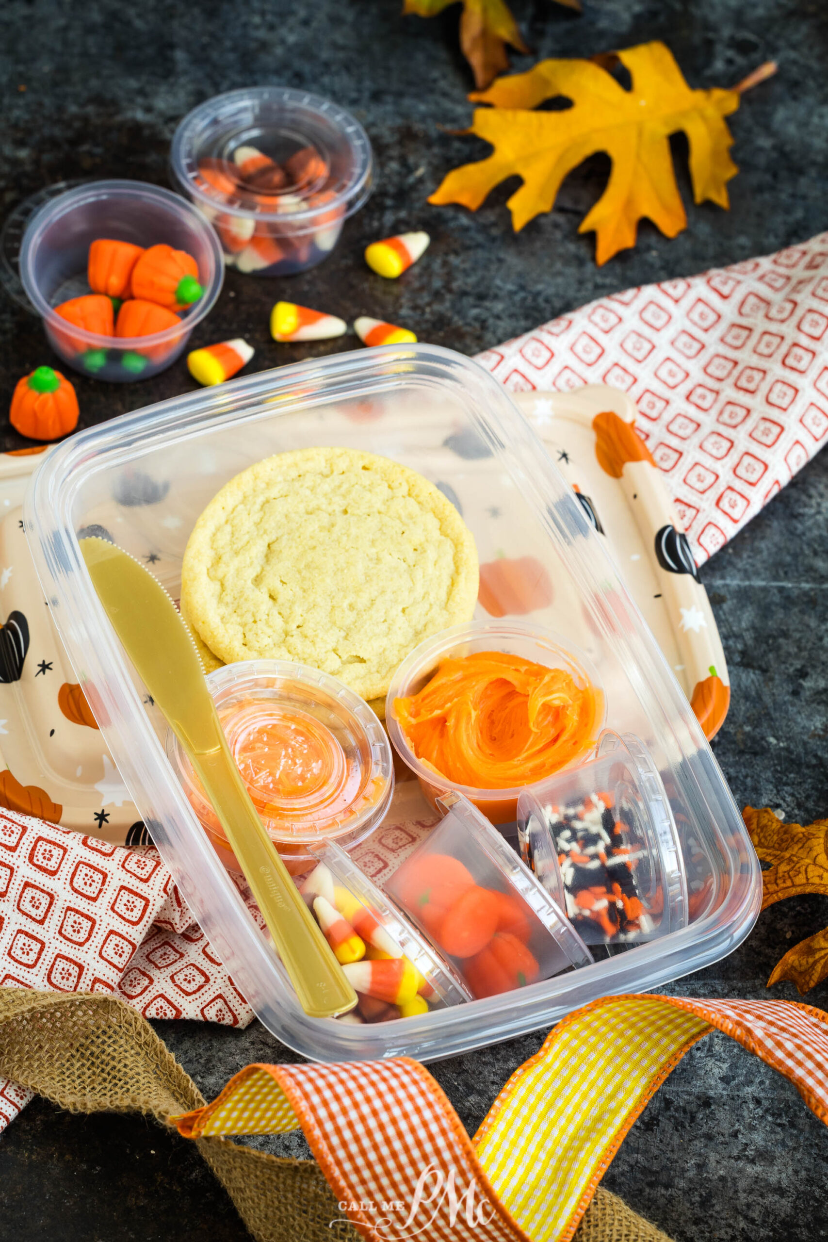 A plastic lunch container holding a cookie, orange icing, candy corn, sprinkles, and gummy pumpkins, with a plastic knife and surrounding fall-themed decorations.