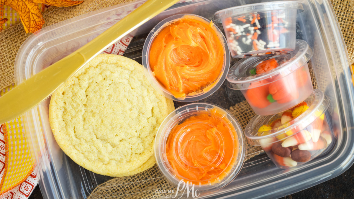 Clear plastic container holding two sugar cookies, two containers of orange frosting, Halloween sprinkles, pumpkin candies, and candy corn.