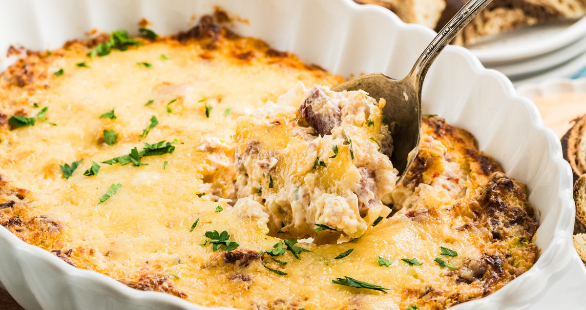 A metal spoon lifting a portion of cheesy, baked reuben dip from a white scalloped baking dish.