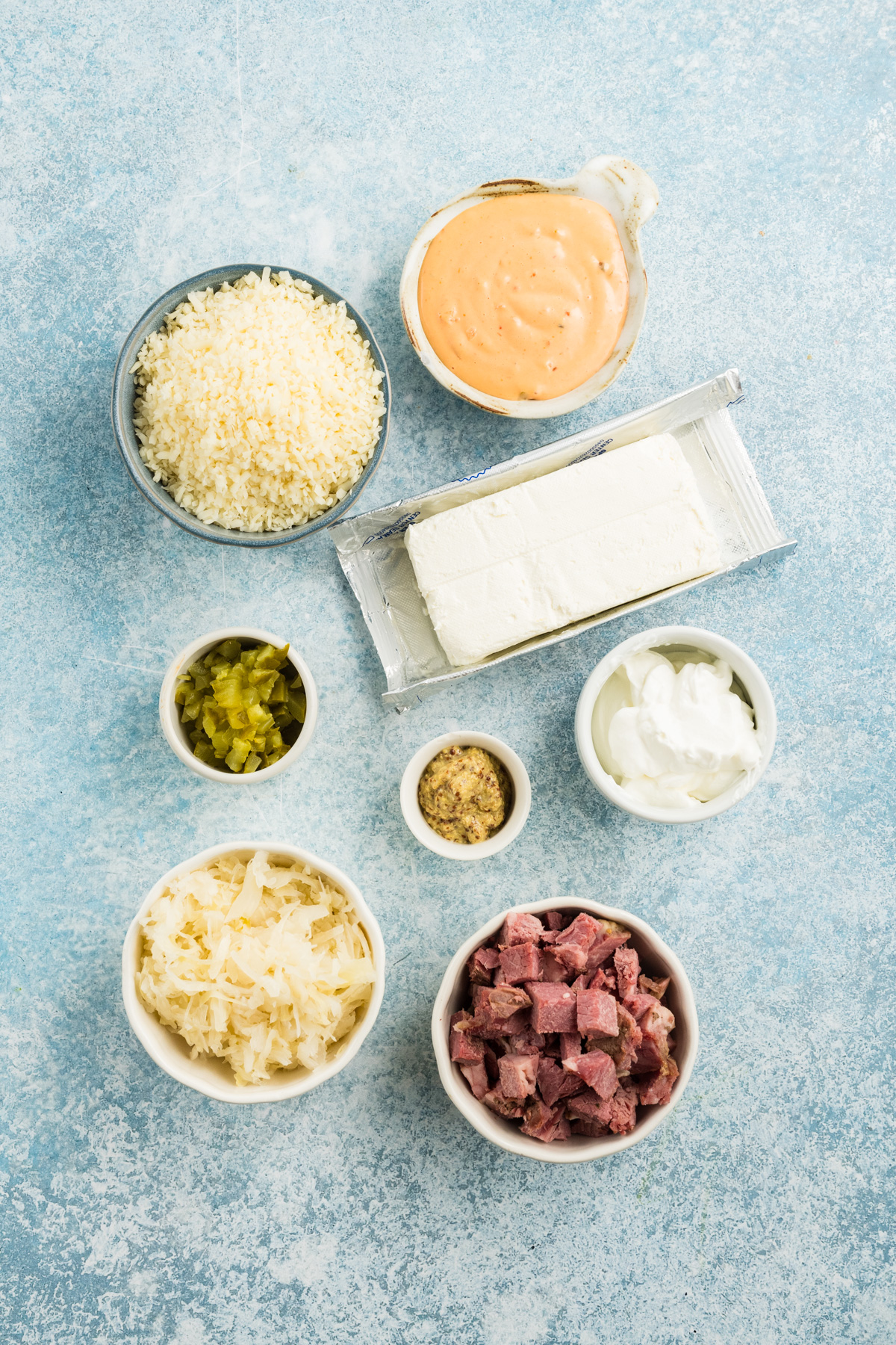 Overhead view of bowls containing shredded cheese, dressing, chopped pickles, mustard, sour cream, sauerkraut, and diced corned beef for a Reuben dip, arranged around a block of cream cheese.