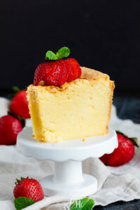 A slice of yellow white chocolate pound cake topped with sliced strawberries and mint, served on a small white pedestal stand.