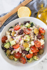 A bowl of Greek salad with cherry tomatoes, cucumbers, olives, red onion, and crumbled feta cheese, with a lemon half and olive oil in the background.
