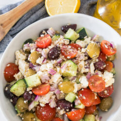 A bowl of Greek salad with cherry tomatoes, cucumbers, olives, red onion, and crumbled feta cheese, with a lemon half and olive oil in the background.