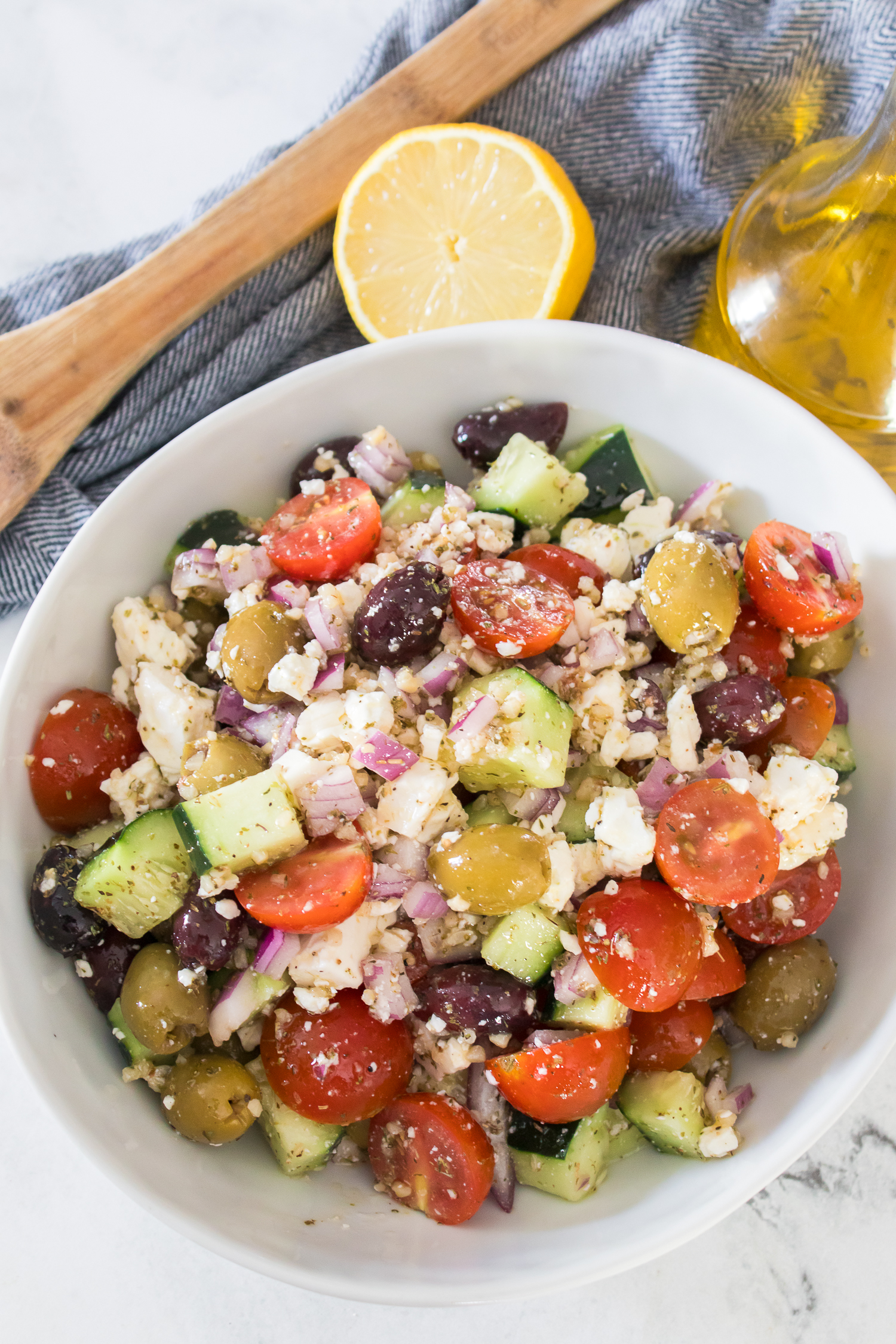 A bowl of vegetables with cherry tomatoes, cucumbers, olives, red onion, and crumbled feta cheese, with a lemon half and olive oil in the background.