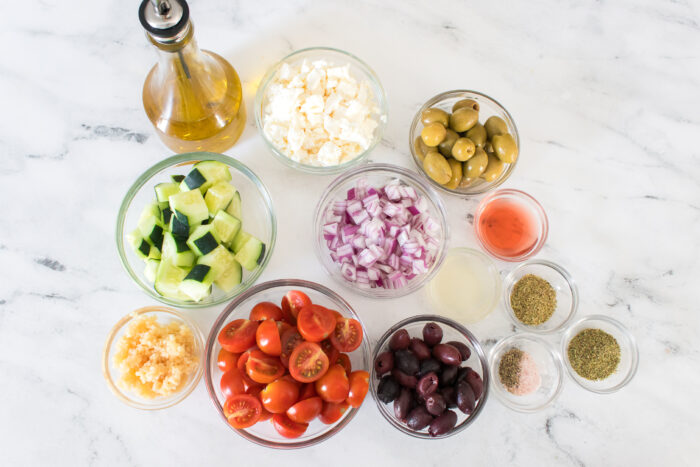 Bowls of chopped cucumber, cherry tomatoes, red onion, feta cheese, green and black olives, minced garlic, herbs, and oil on a white marble surface.