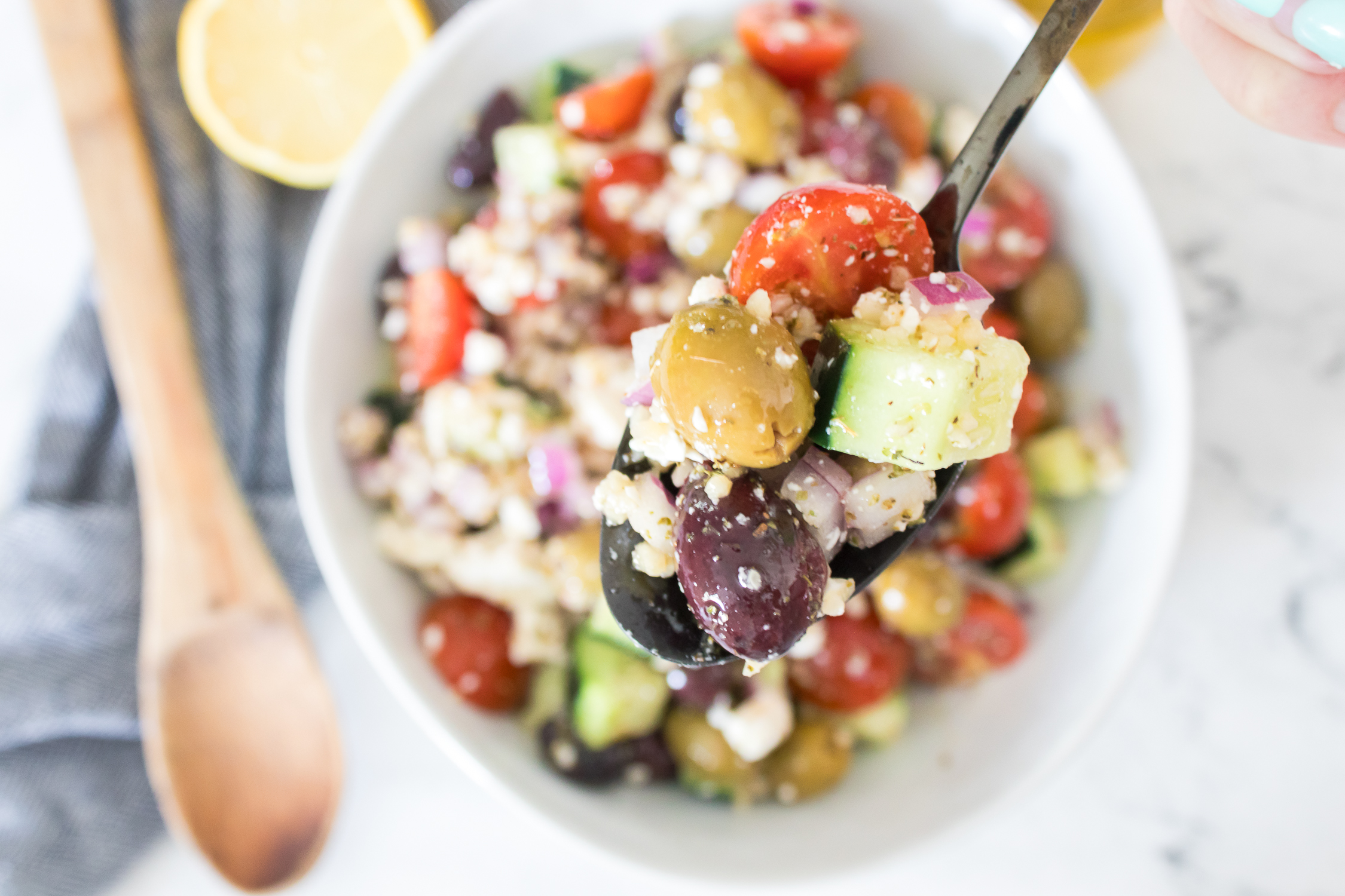 A spoonful of Greek salad containing olives, cucumber, cherry tomatoes, red onion, and crumbled cheese, held over a bowl of the same salad.