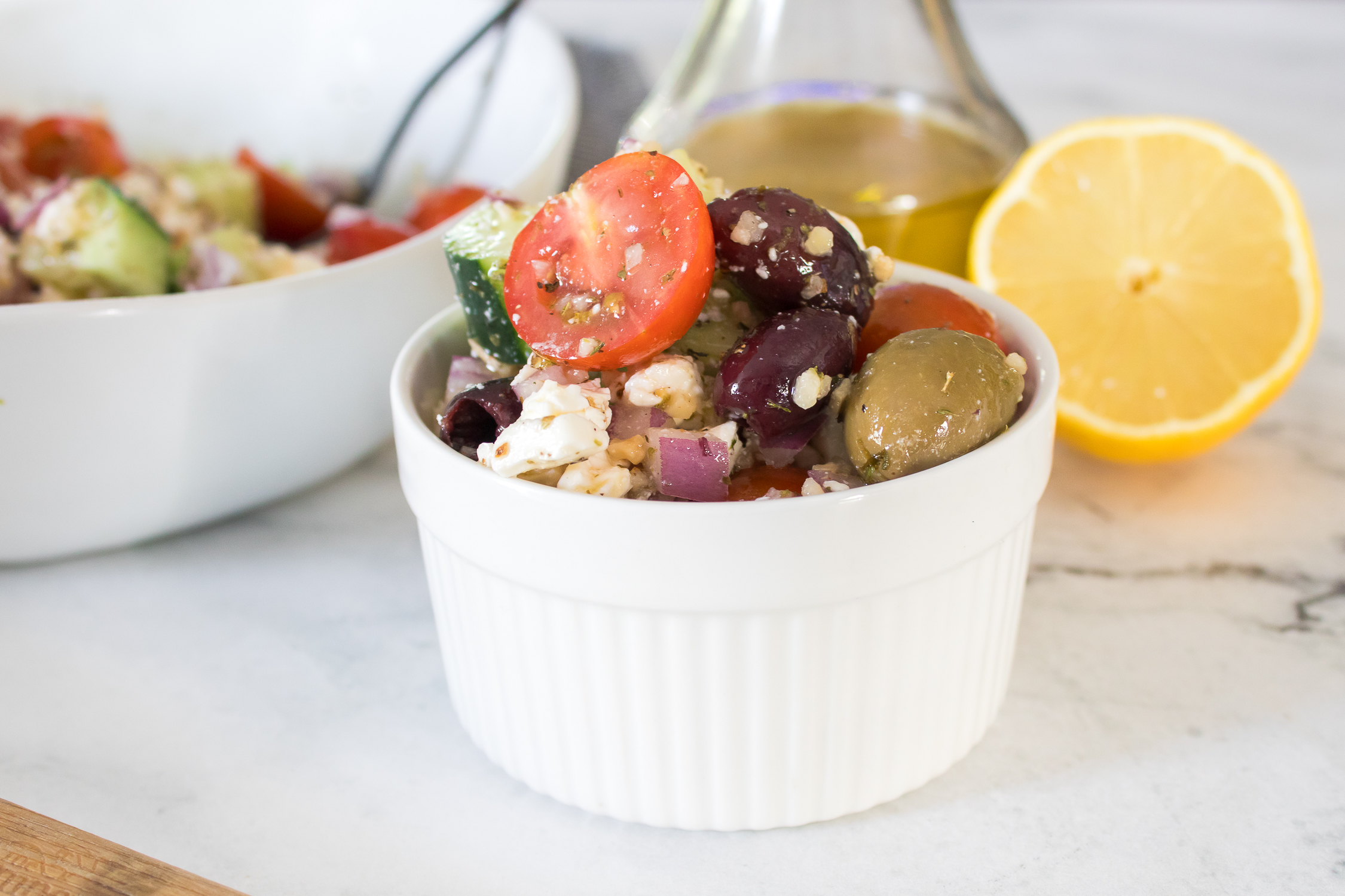 A white ramekin filled with Greek salad containing cherry tomatoes, olives, cucumber, red onion, and feta cheese, with a lemon half and a bottle of olive oil nearby.