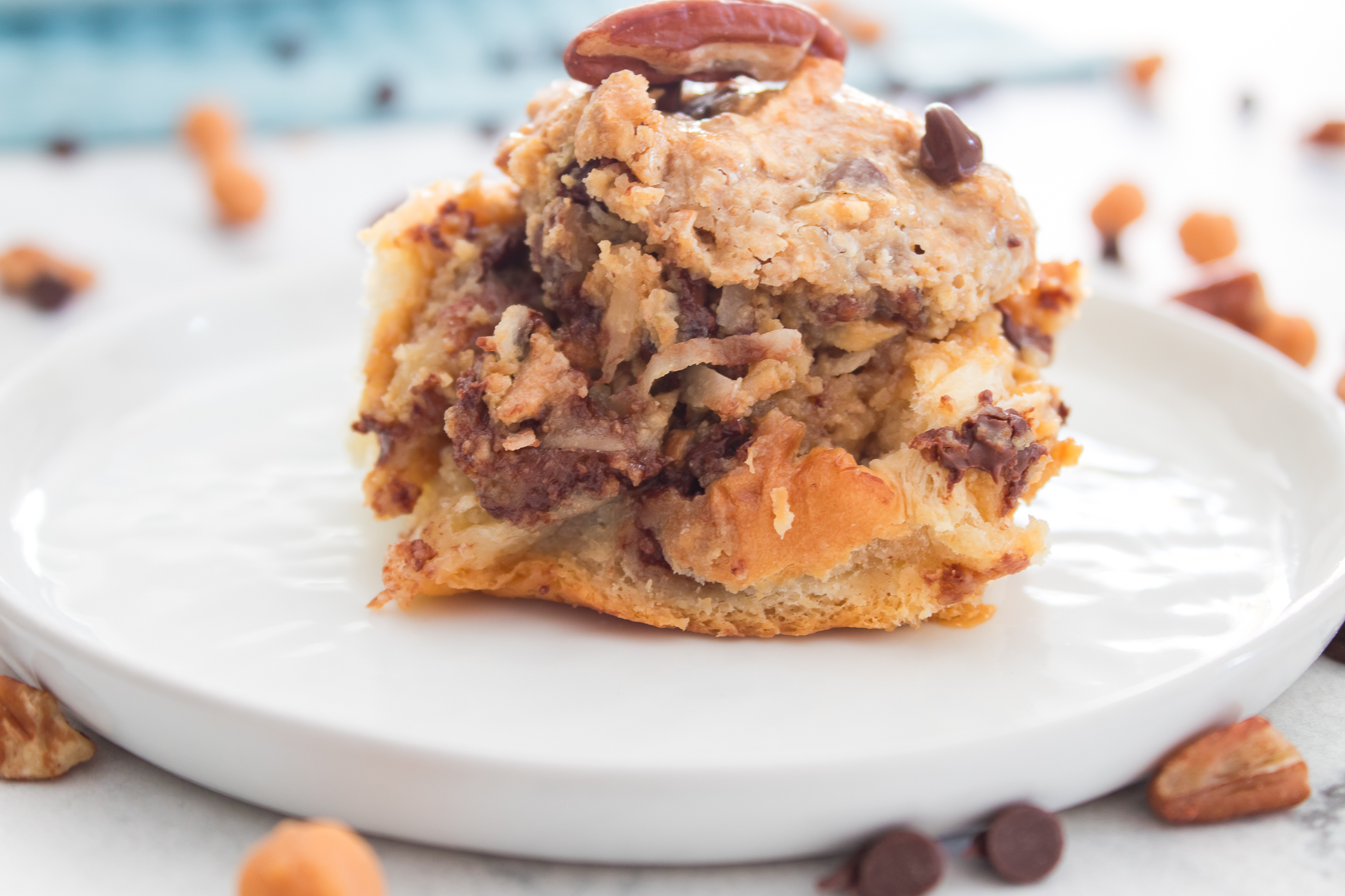 A serving of Texas Trash sticky buns topped with a pecan on a white plate, surrounded by scattered chocolate chips and pecans.